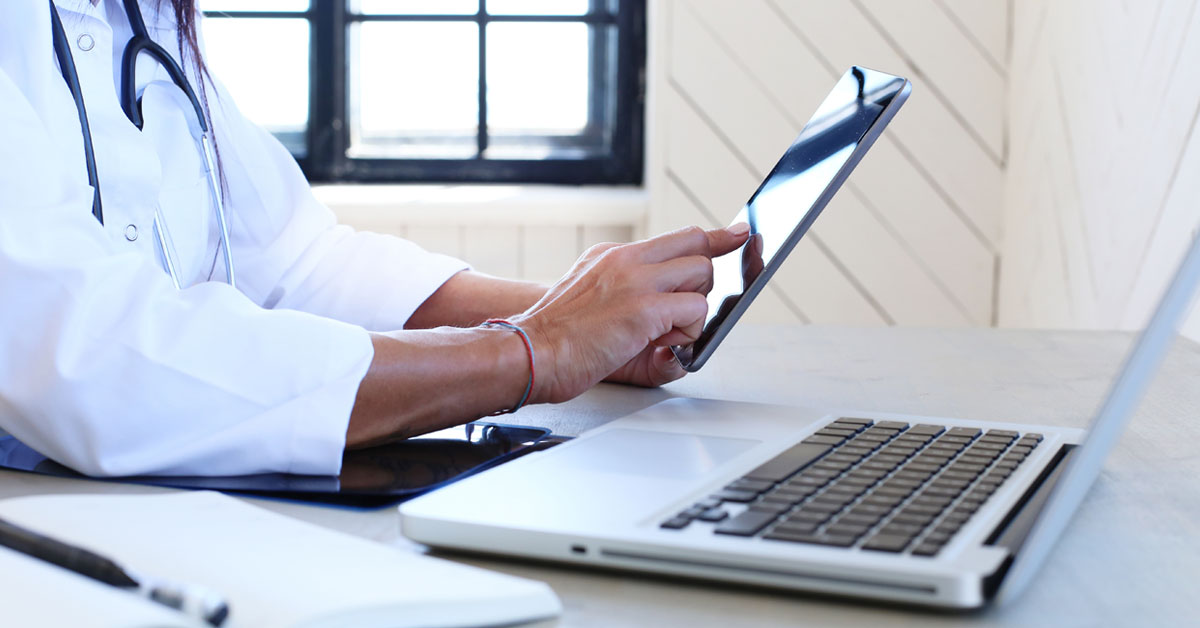 Un medico sta utilizzando un tablet, con un laptop aperto e una penna posizionata lì accanto su un foglio sul tavolo. Può rappresentare la determinazione del personale sanitario nell'affrontare le sfide lanciate alla sanità dalla digitalizzazione.